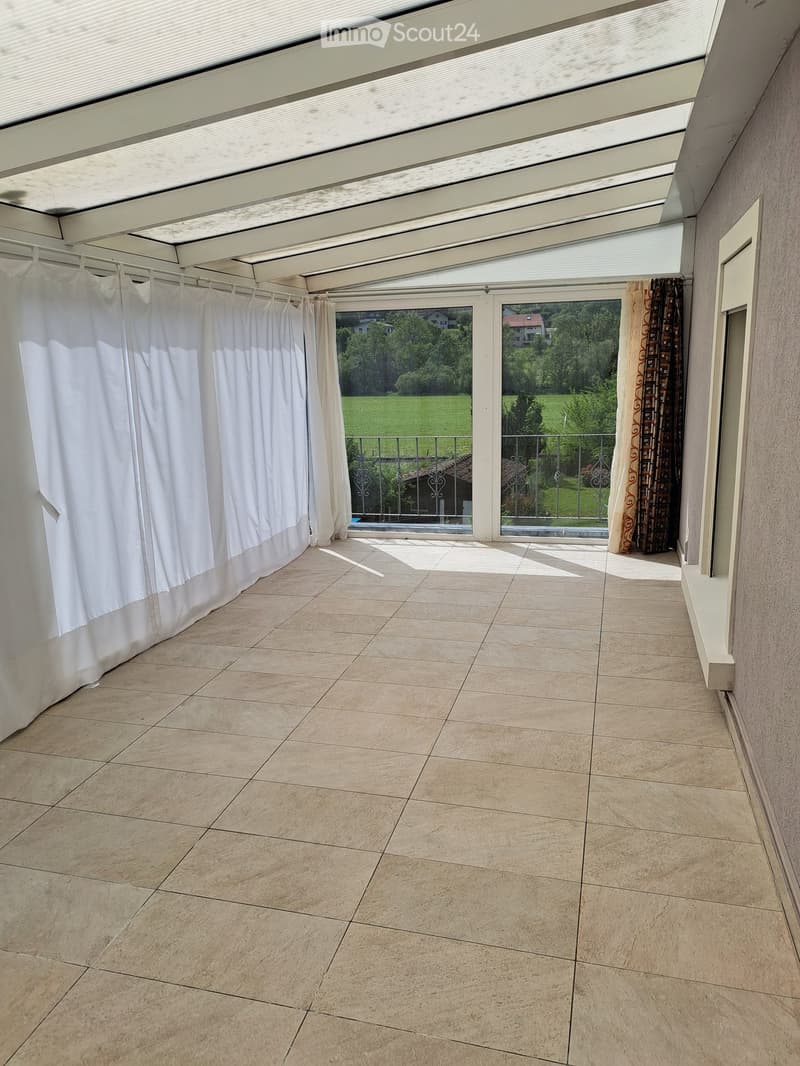 Covered balcony with tiled floor, white curtains, and large windows offering a view of a green landscape.