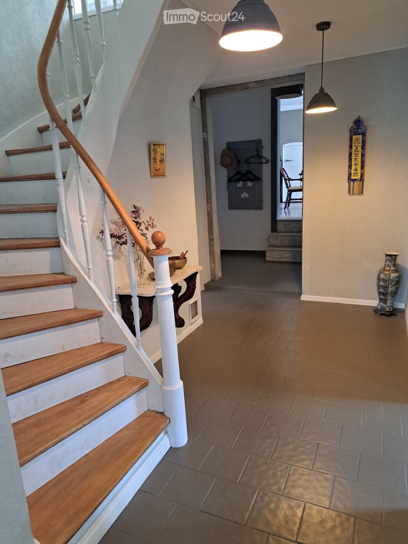 Spacious hallway with wooden stairs, white balustrade, pendant lights, and a small console table with a vase of flowers. A doorway leads to another room with a visible chair and coat rack.