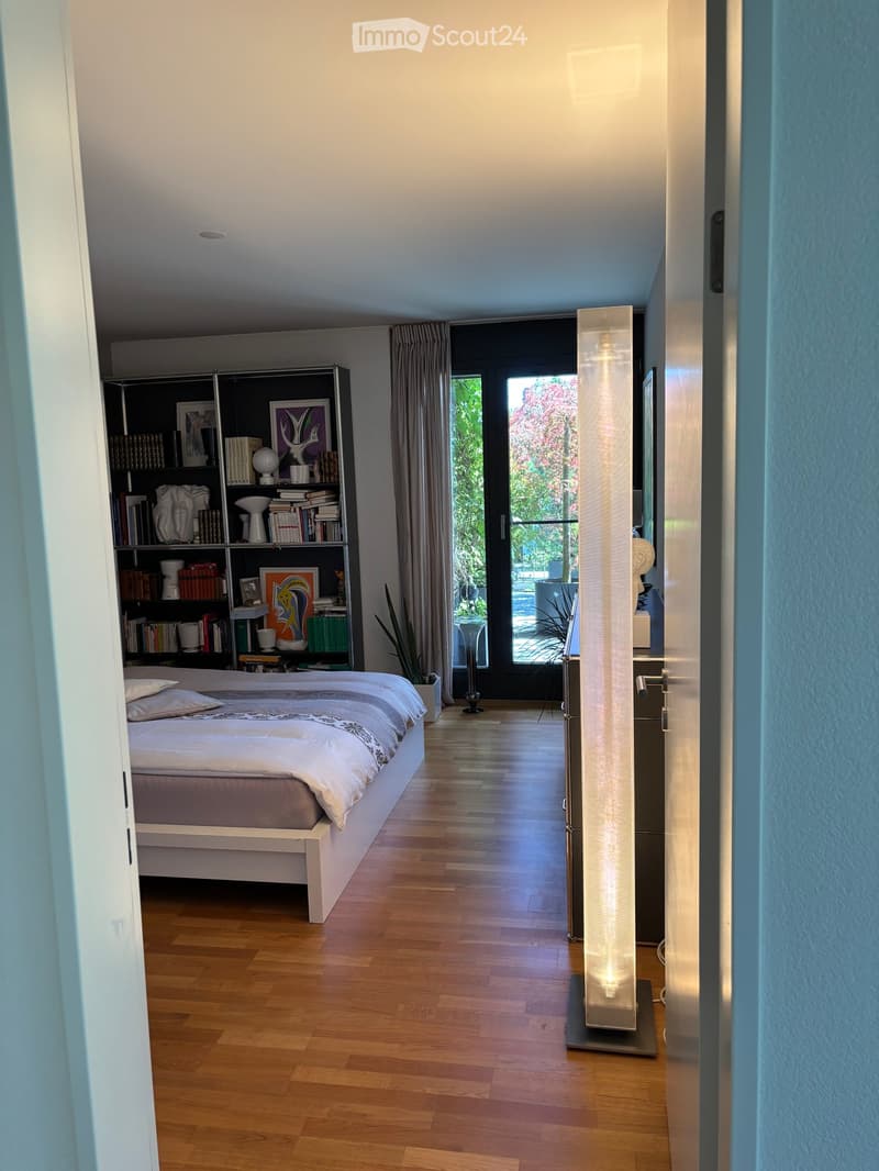 Modern bedroom with a white bed, wooden floor, bookshelf, and large window with curtain leading to a balcony.