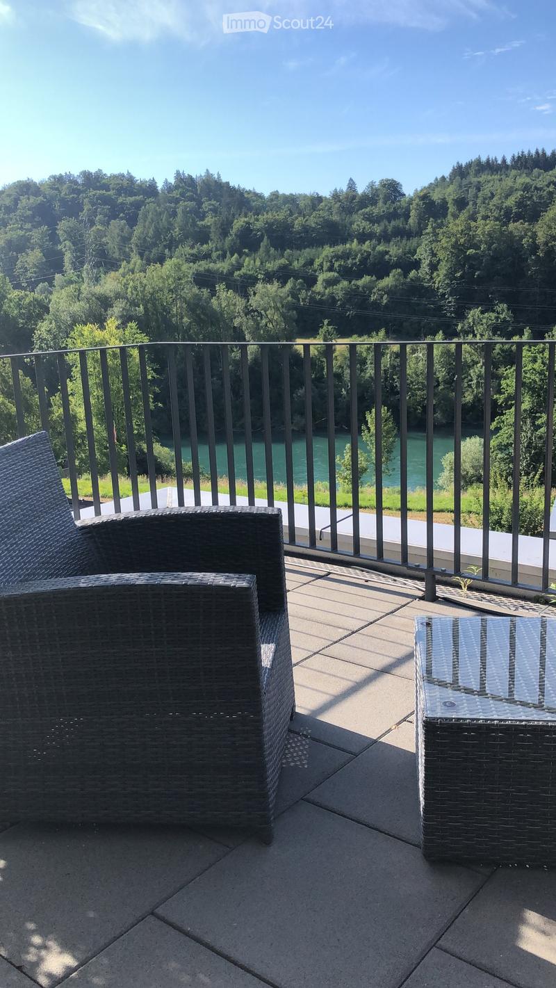 Balcony with a view of a river and forested area, featuring a wicker chair and a small table.