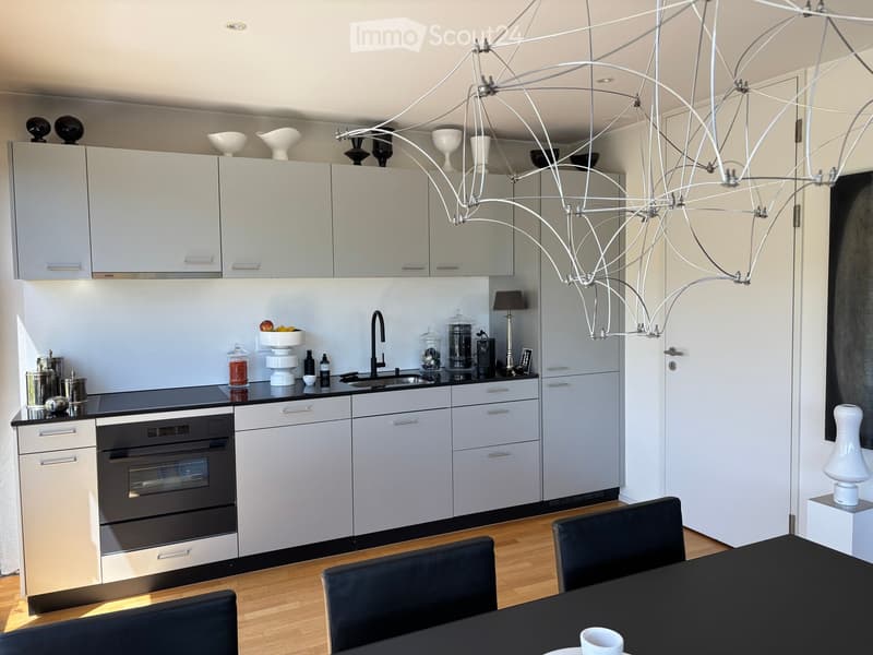 Modern kitchen with grey cabinets, black countertop, stainless steel sink, built-in oven, and a black dining table with chairs.