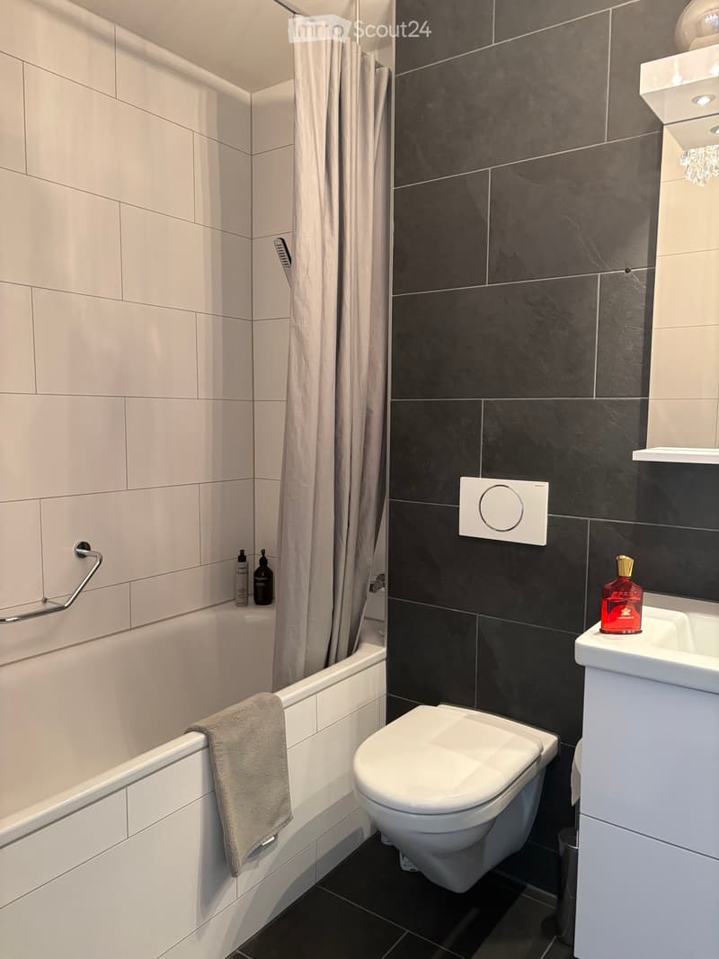 Modern bathroom with a bathtub, toilet, sink, and a towel draped over the edge of the tub. The walls are tiled in white and dark gray, and there is a shower curtain in the background.