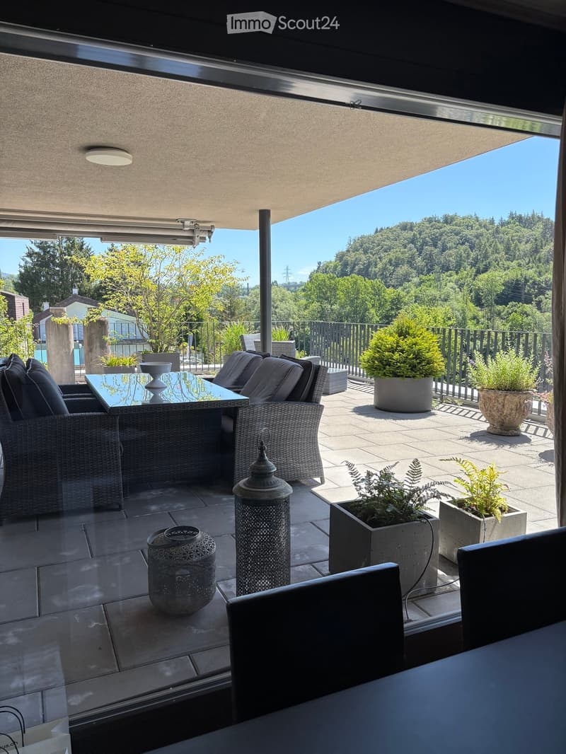 Balcony with a modern design, featuring a glass roof, outdoor furniture including a table and chairs, decorative lanterns, and potted plants, overlooking a lush green landscape.