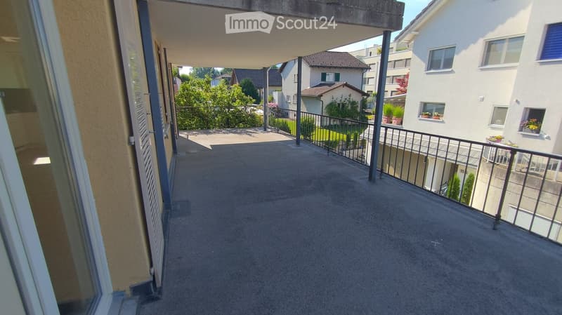Balcony with concrete floor, metal railing, overlooking a residential area with houses and greenery.