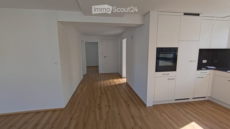A narrow hallway with wooden flooring and white walls, leading to a kitchen area with white cabinets and a built-in oven.