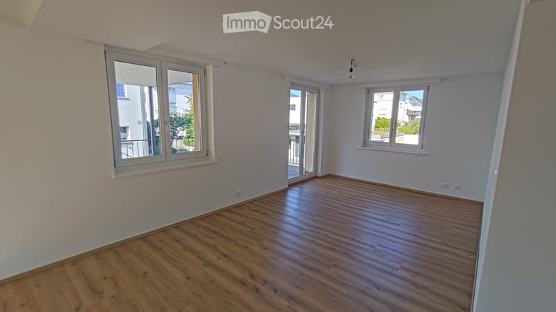 Empty room with wooden flooring, white walls, and large windows with white frames. The room has a sliding glass door leading to a balcony area.