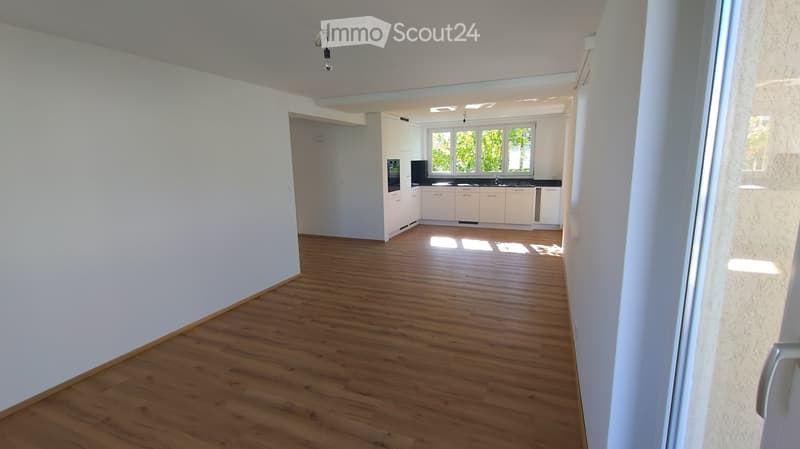 Empty room with wooden flooring and white walls, leading to a small kitchen area with white cabinets and black countertops.