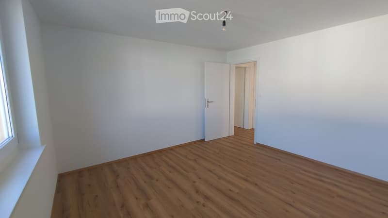 Empty room with light wooden flooring, white walls, and a single door leading to another space. Natural light comes through a window on the left side.