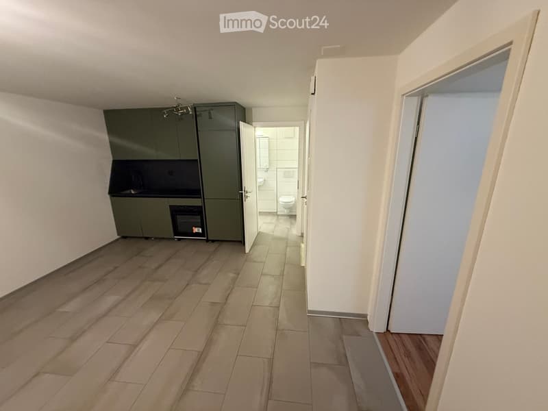 Empty hallway with tiled floor, white walls, and a small kitchen area with dark green cabinets and a sink, leading to a bathroom with a toilet and sink.