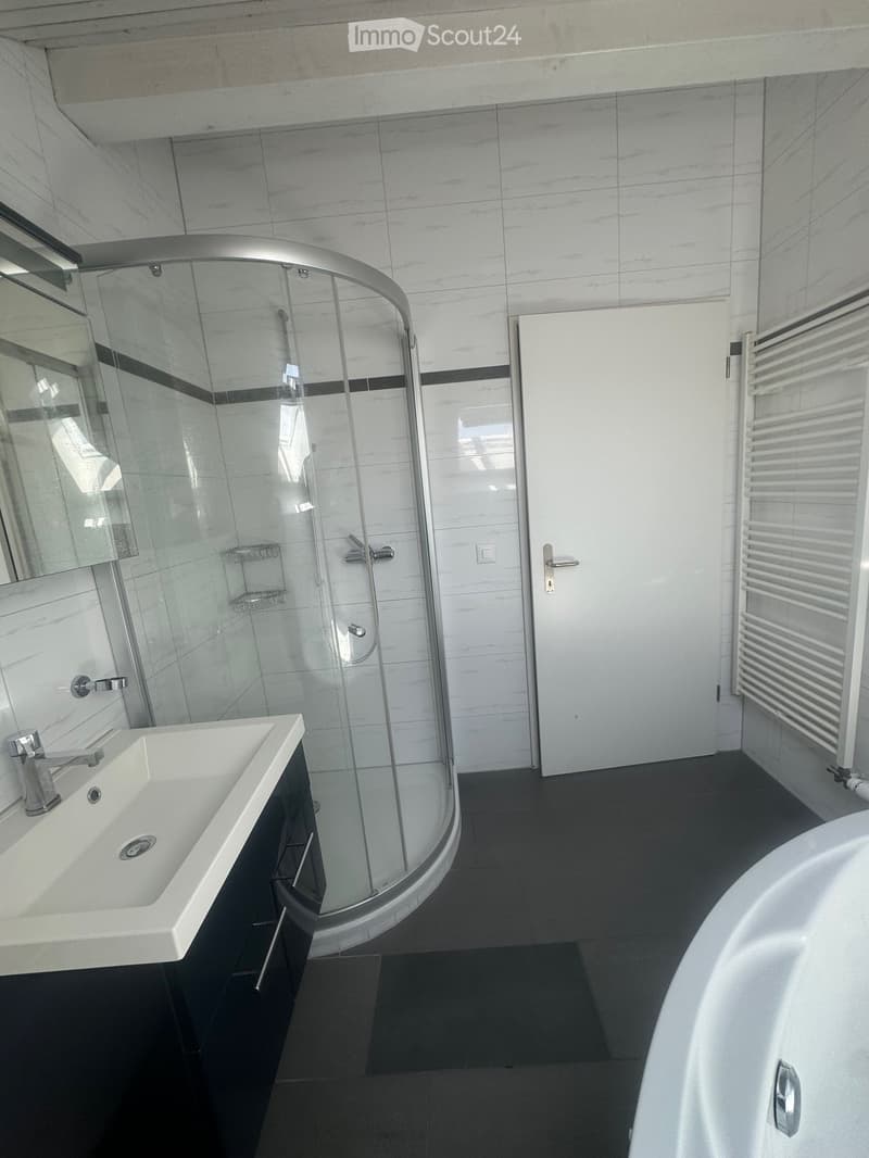 Modern bathroom with a white sink, chrome faucet, bathtub, glass-enclosed shower, and a radiator. The walls are tiled in a light marble pattern, and the floor is dark grey.