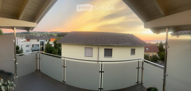 Balcony with modern glass railing, overlooking a residential area with houses and a sunset view.