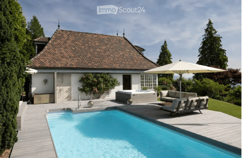 A modern outdoor area features a swimming pool, a hot tub, lounge chairs, and a large umbrella providing shade. The area is surrounded by lush greenery and a well-maintained lawn.