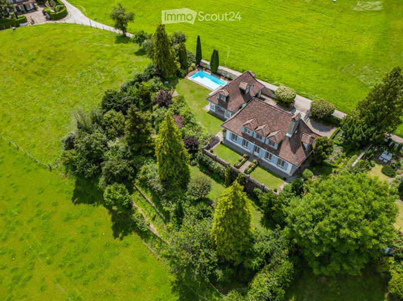 Aerial view of a large, well-maintained garden surrounding a two-story house with a red-tiled roof and white walls. The garden features lush green lawns, mature trees, and a swimming pool near the house.