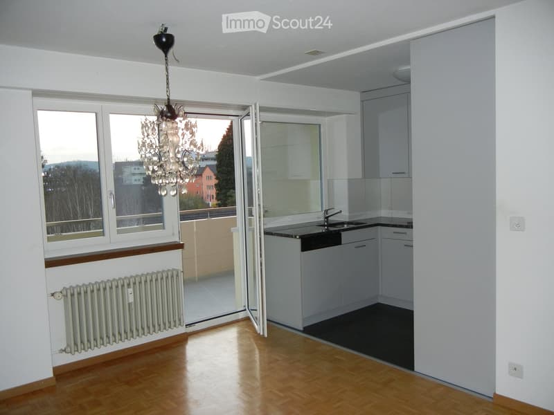 Bright, modern living space with wooden flooring, large windows, a chandelier, radiator, and a small kitchen area with white cabinets and a sink.
