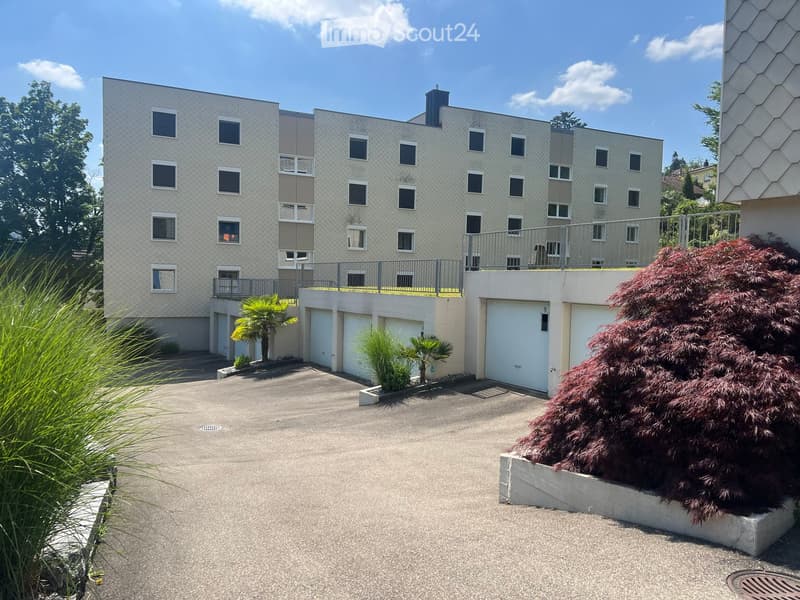 Modern multi-story building with balconies, garage, and landscaped surroundings including palm trees and shrubs.