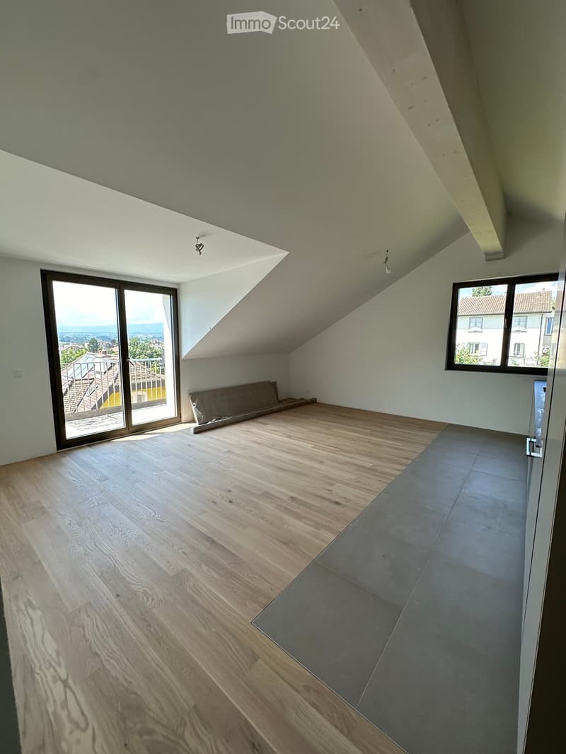 Empty attic room with wooden flooring, large windows, and a view of the cityscape.