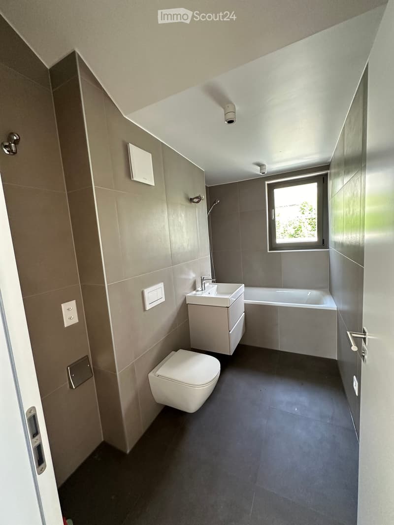 Modern bathroom with a toilet, sink, bathtub, and a window letting in natural light. The walls are tiled in neutral tones, and the floor is dark grey.