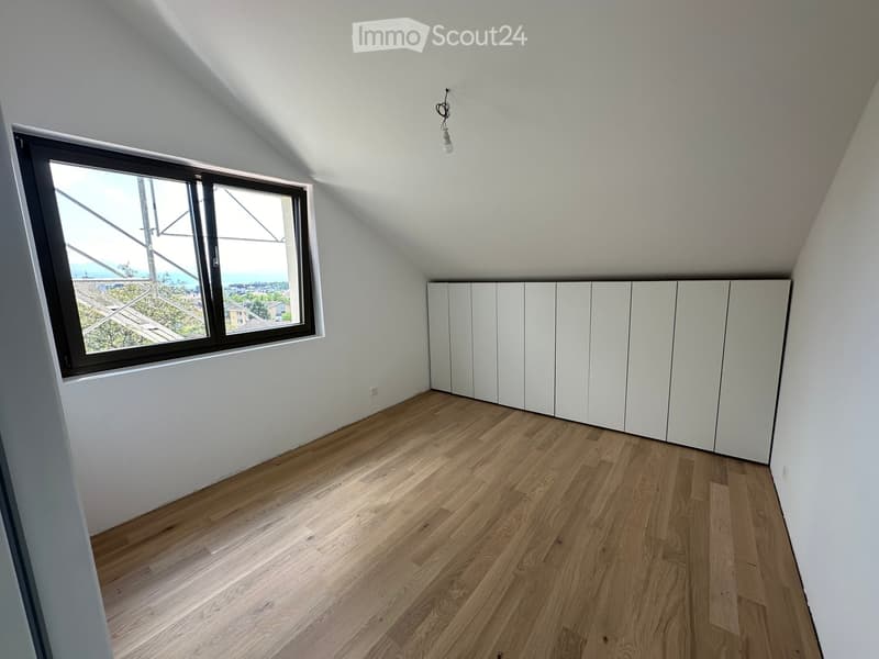 Empty attic room with wooden flooring, white walls, built-in white cabinets, and a large window offering a view of the city and sea.