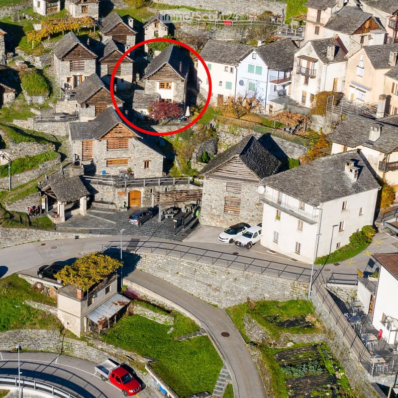 Aerial view of a small village with stone houses, narrow roads, and parked cars. The houses have traditional stone facades and wooden shutters, with some featuring balconies and terraces.