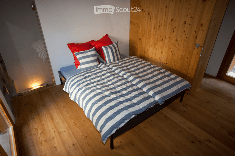 Cozy bedroom with a wooden floor, a bed with blue and white striped bedding and red pillows, a nightstand with a lamp, and a wooden wall panel.