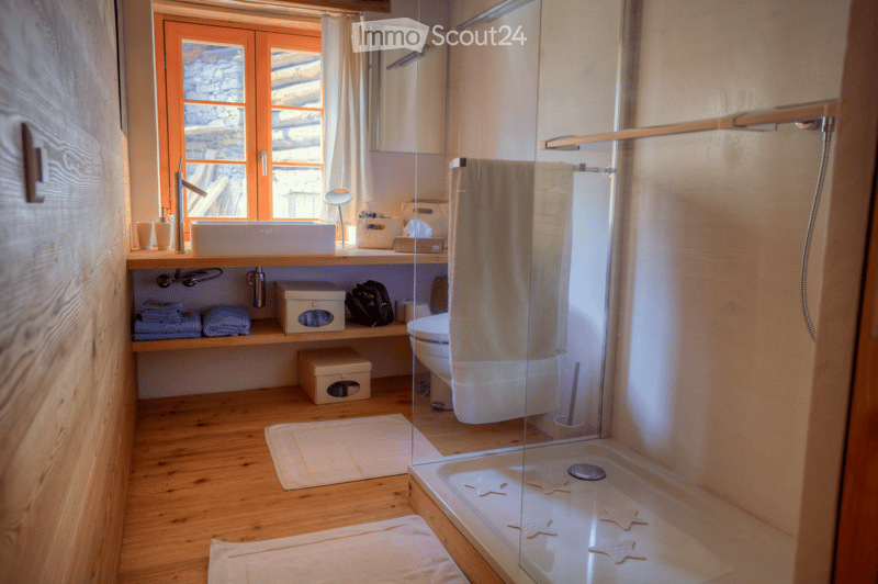 Modern bathroom with wooden flooring, a large sink with a modern faucet, a mirror, a toilet, a glass-enclosed shower with star-shaped floor tiles, and neatly arranged towels and storage boxes.