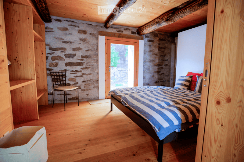 Cozy bedroom with wooden floors, exposed wooden beams, stone walls, a bed with striped bedding, a chair, and built-in wooden shelves.