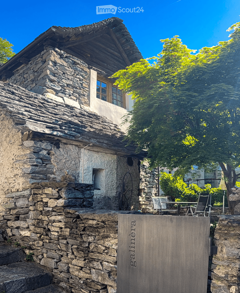A rustic stone house with a steep, multi-level roof and wooden-framed windows, surrounded by lush greenery and a small outdoor seating area with chairs and a table.
