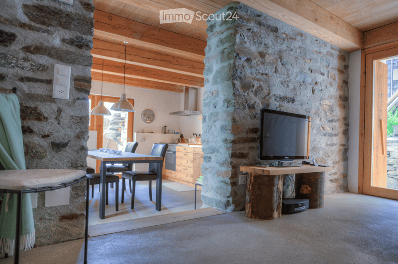 Cozy living space with exposed wooden beams, stone walls, and a mix of modern and rustic furniture including a dining table, chairs, TV, and wooden cabinet.