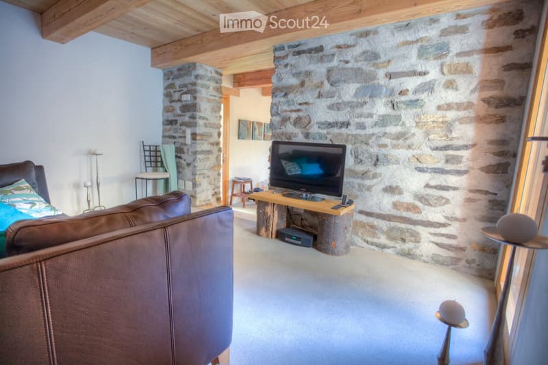 Living room with exposed wooden beams, stone walls, a brown leather sofa, a wooden TV stand, and a small round table with a lamp.
