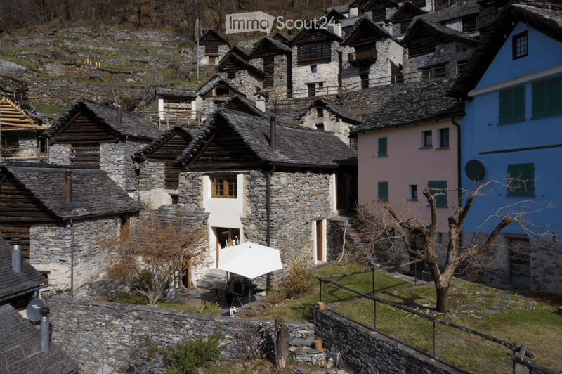 A picturesque village with stone houses, wooden beams, and colorful shutters, surrounded by a stone wall and greenery.