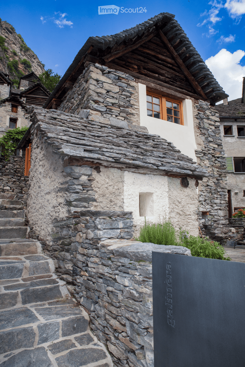 Traditional stone house with a steep, wooden roof and multiple windows, surrounded by a stone pathway and small garden area.