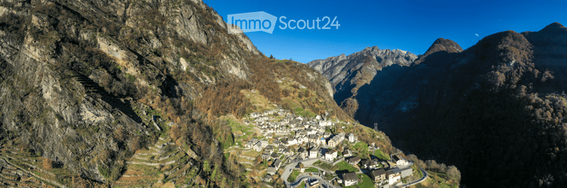A small village nestled in a mountainous valley, surrounded by rocky cliffs and autumn-colored trees. The village consists of white stone houses with dark roofs, arranged along winding roads and pathways.