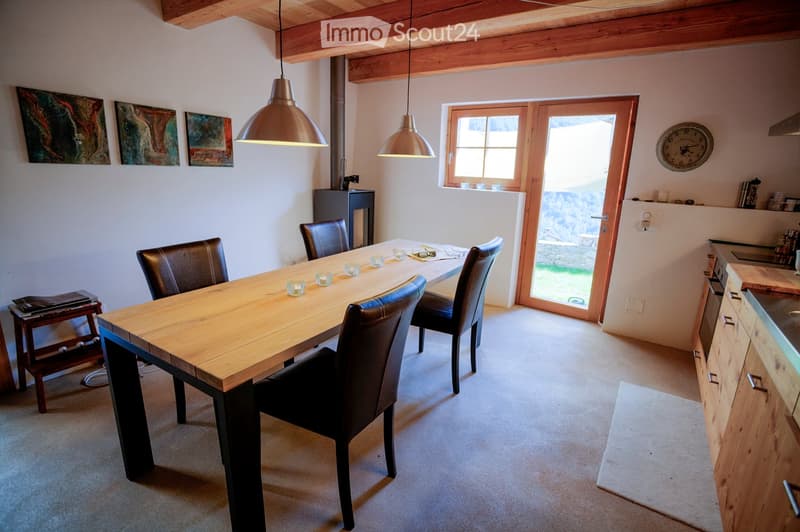 Dining room with a long wooden dining table, four black leather chairs, pendant lights, a clock, and a small wooden stool with books.