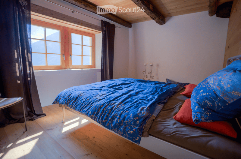 Cozy bedroom with wooden floors, exposed wooden beams, large window with dark curtains, and a neatly made bed with blue bedding and pillows.