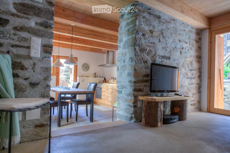 A cozy living and dining area with exposed wooden beams, stone walls, and a mix of modern and rustic furniture, including a dining table, chairs, a TV, and a wooden cabinet.