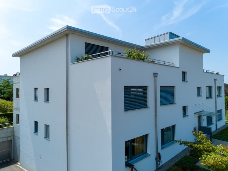 Modern two-story house with white exterior, multiple windows, balconies, and greenery around the base.