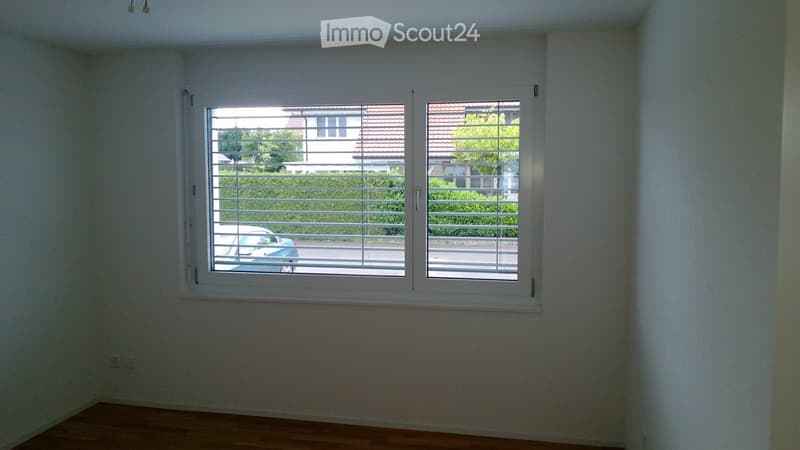 Empty room with light-colored walls and wooden flooring, featuring two large windows with white frames and blinds.