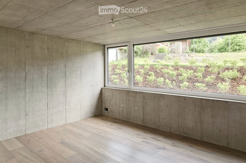 Minimalist unfurnished room with exposed wooden ceiling, concrete walls, and large window overlooking a landscaped garden.