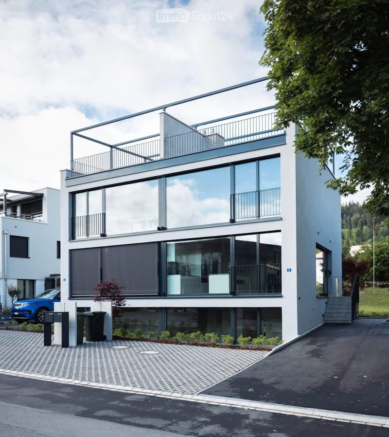 Modern two-story house with large glass windows, balconies, and a raised ground floor. Features a paved driveway, small garden area, and a blue car parked nearby.