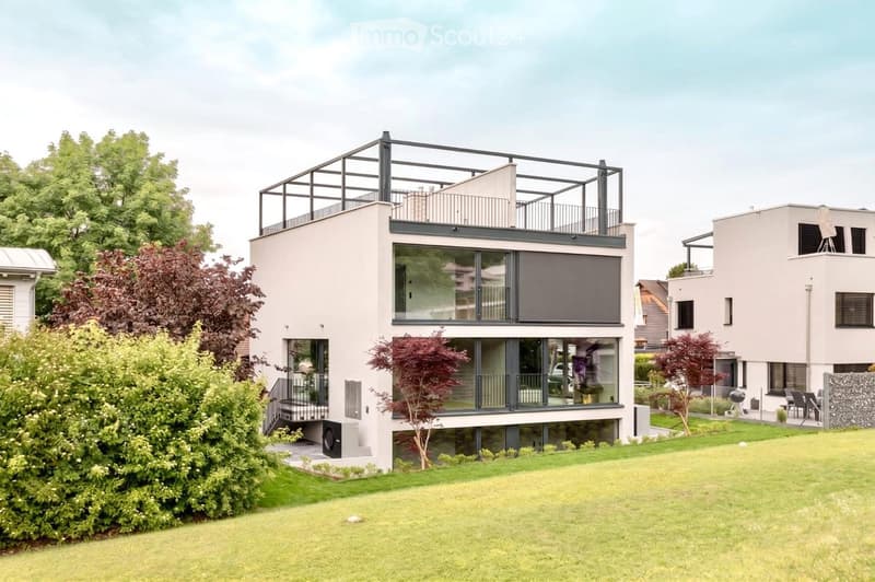 Modern two-story house with large glass windows, a raised ground floor, and a rooftop terrace with a metal railing.