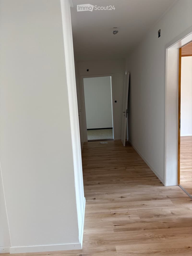 Empty hallway with light wooden flooring and white walls, featuring a partially open door leading to another room.