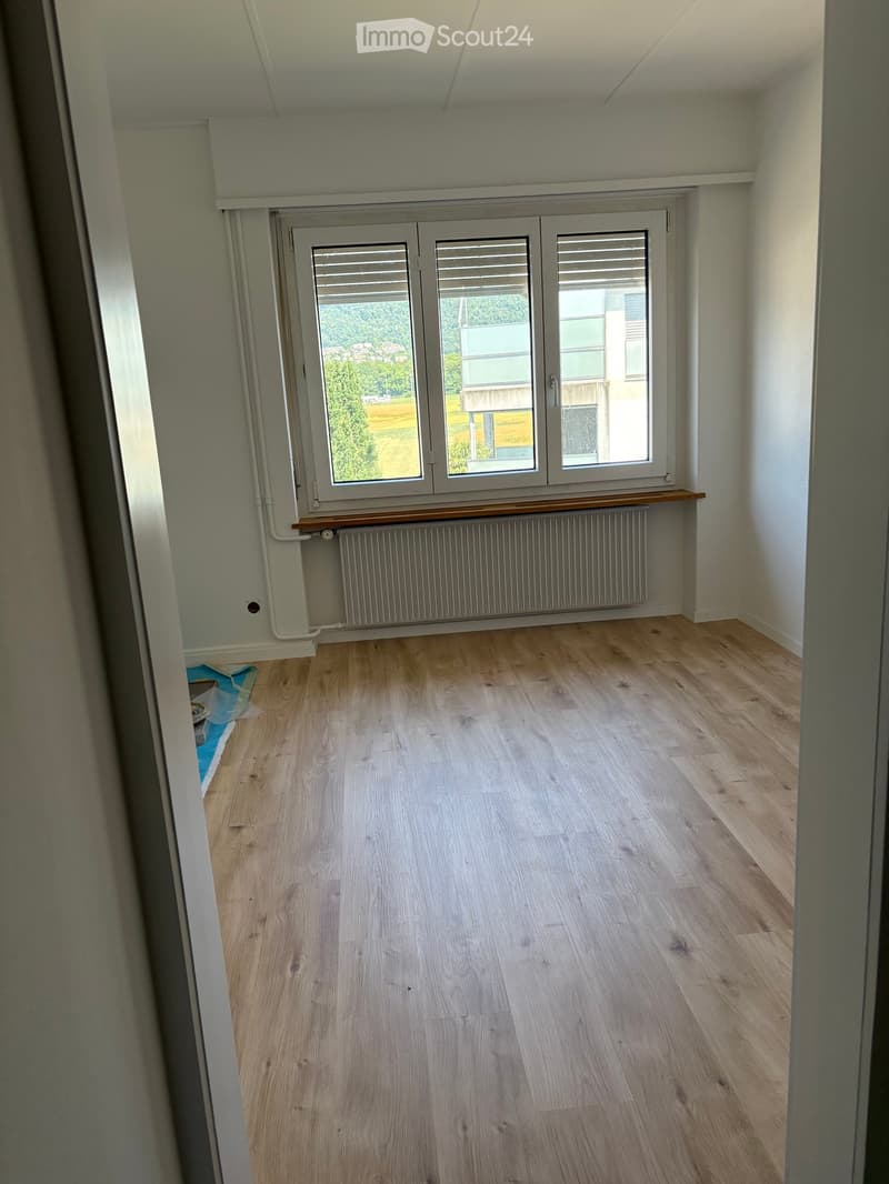 Empty room with light wooden flooring, white walls, and large windows with blinds. A radiator is visible below the windows.