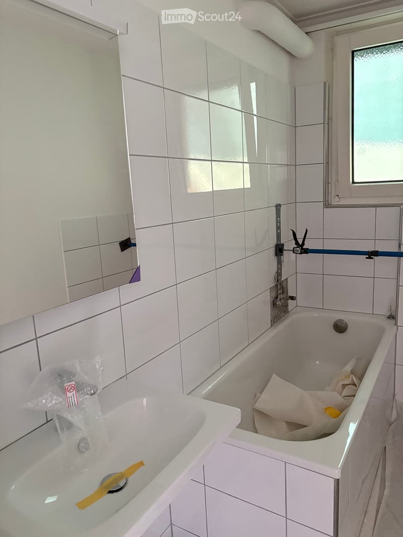Bathroom with white tiled walls and floor, a bathtub, a mirror, and a window with frosted glass. There is a towel inside the bathtub and some plastic bags on the floor.