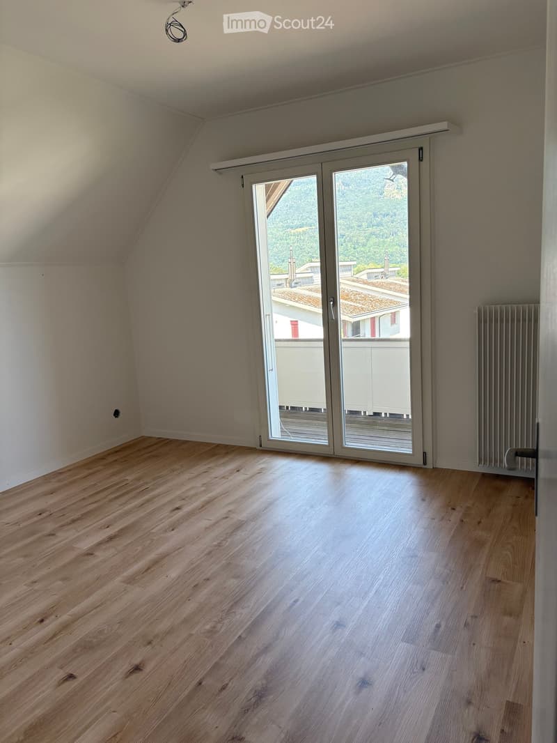Empty room with wooden flooring, large sliding glass door leading to a balcony, radiator on the wall, and exposed wooden beams on the ceiling.