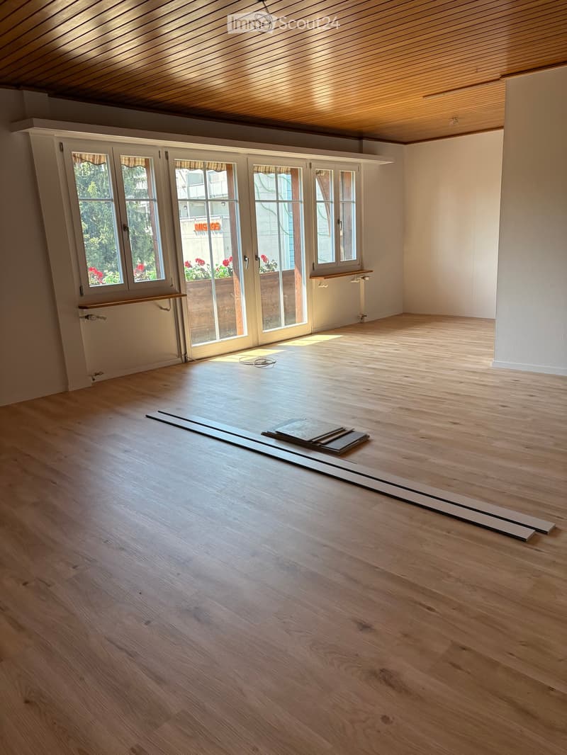 Empty room with wooden flooring, white walls, and large windows with blinds. Wooden beams on the ceiling and a small shelf on one wall.