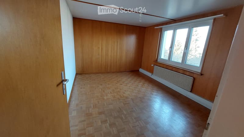 Empty room with wooden flooring, wooden walls, large window, radiator, and a door with a silver handle.