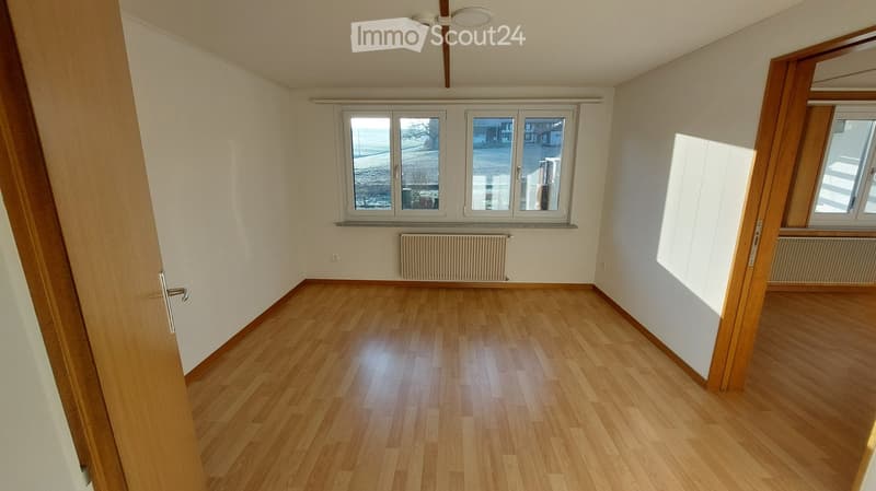 Empty room with wooden flooring, white walls, and large windows offering a view of the street. Radiators are visible under the windows.