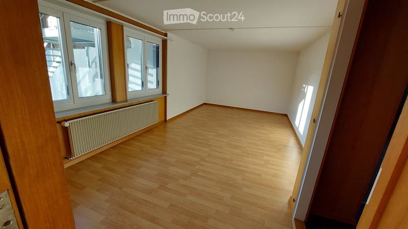 Empty room with wooden flooring, white walls, and large windows with wooden frames. A radiator is visible under one window.