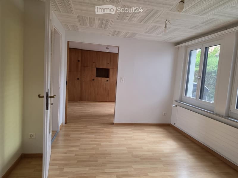 Empty room with light wooden flooring, white walls, large windows, and a wooden cabinet in the background.