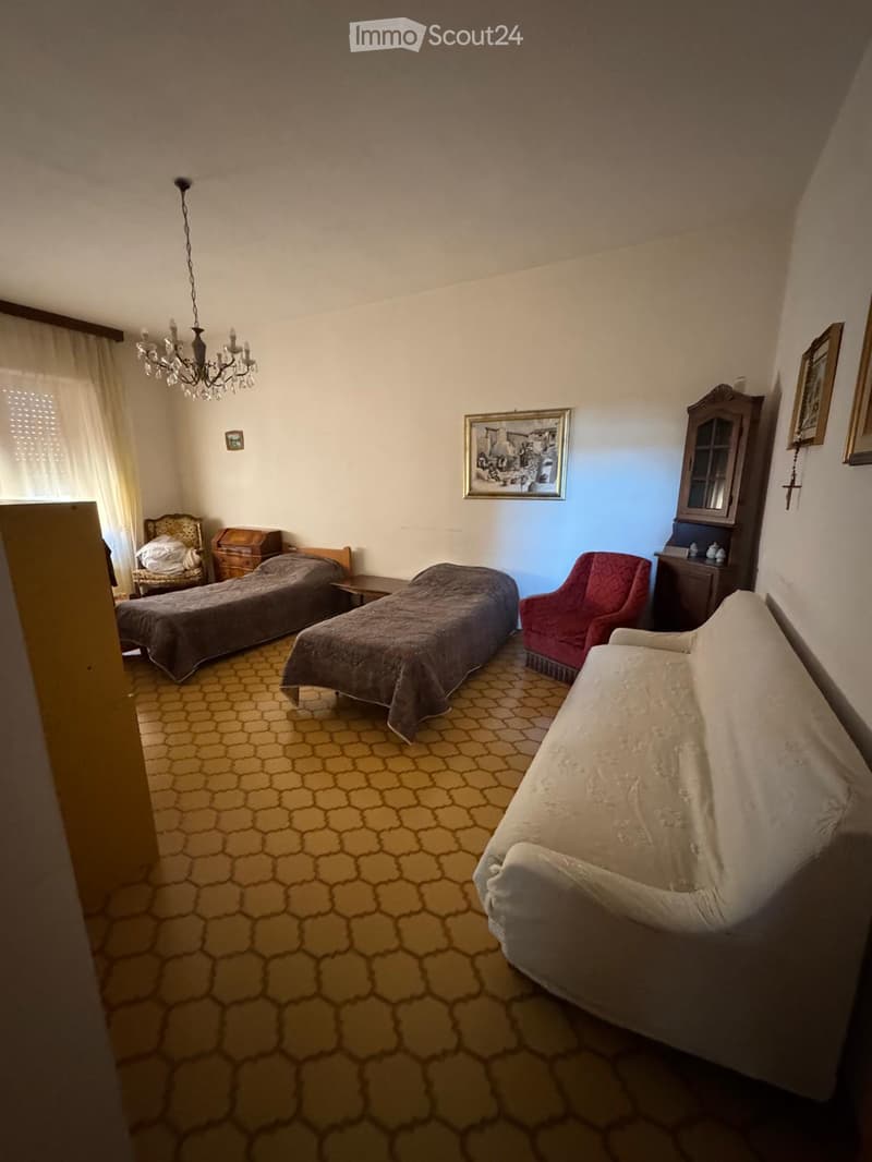 A furnished room with a sofa, two beds, a chair, a small table, a wooden cabinet, and a chandelier.
